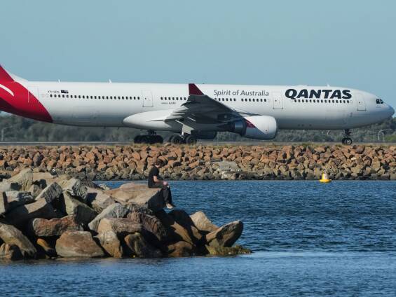 A fisherman sits on the edge of a rock wall on Botany Bay as a Qantas passenger jet prepares for take off from Sydney Airport. Picture by AP/Mark Baker